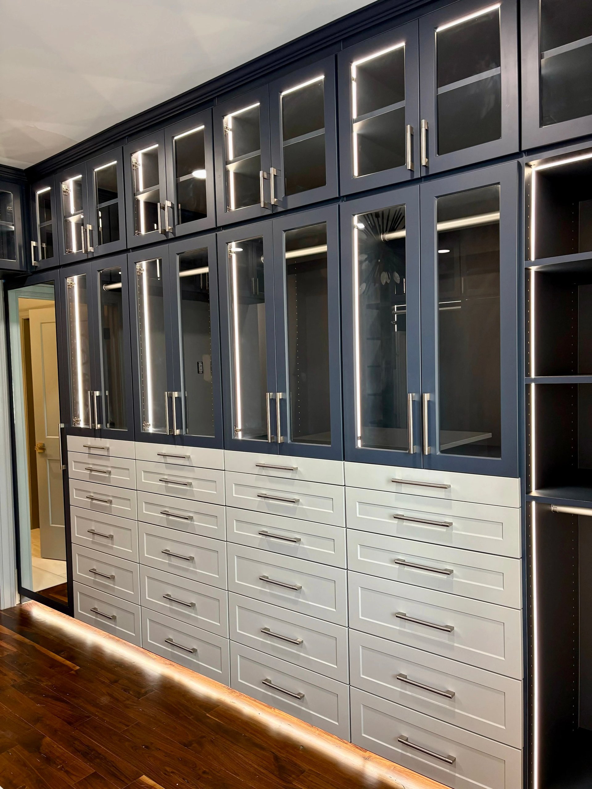 Modern walk-in closet with dark blue glass-front upper cabinets and white lower drawers, illuminated by LED lighting