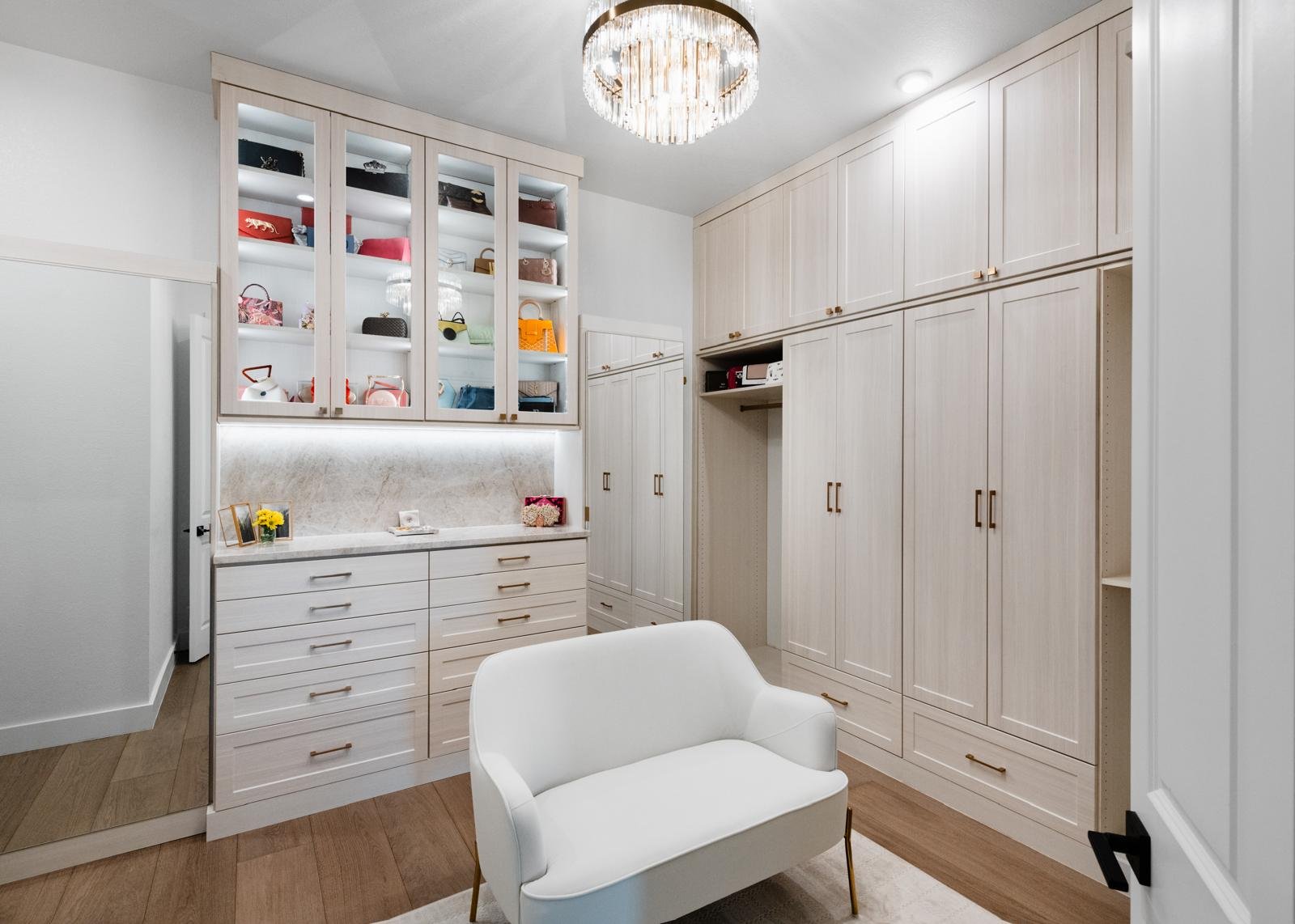 Modern white walk-in closet with built-in shelving, white armchair, wooden flooring, and crystal ceiling light fixture