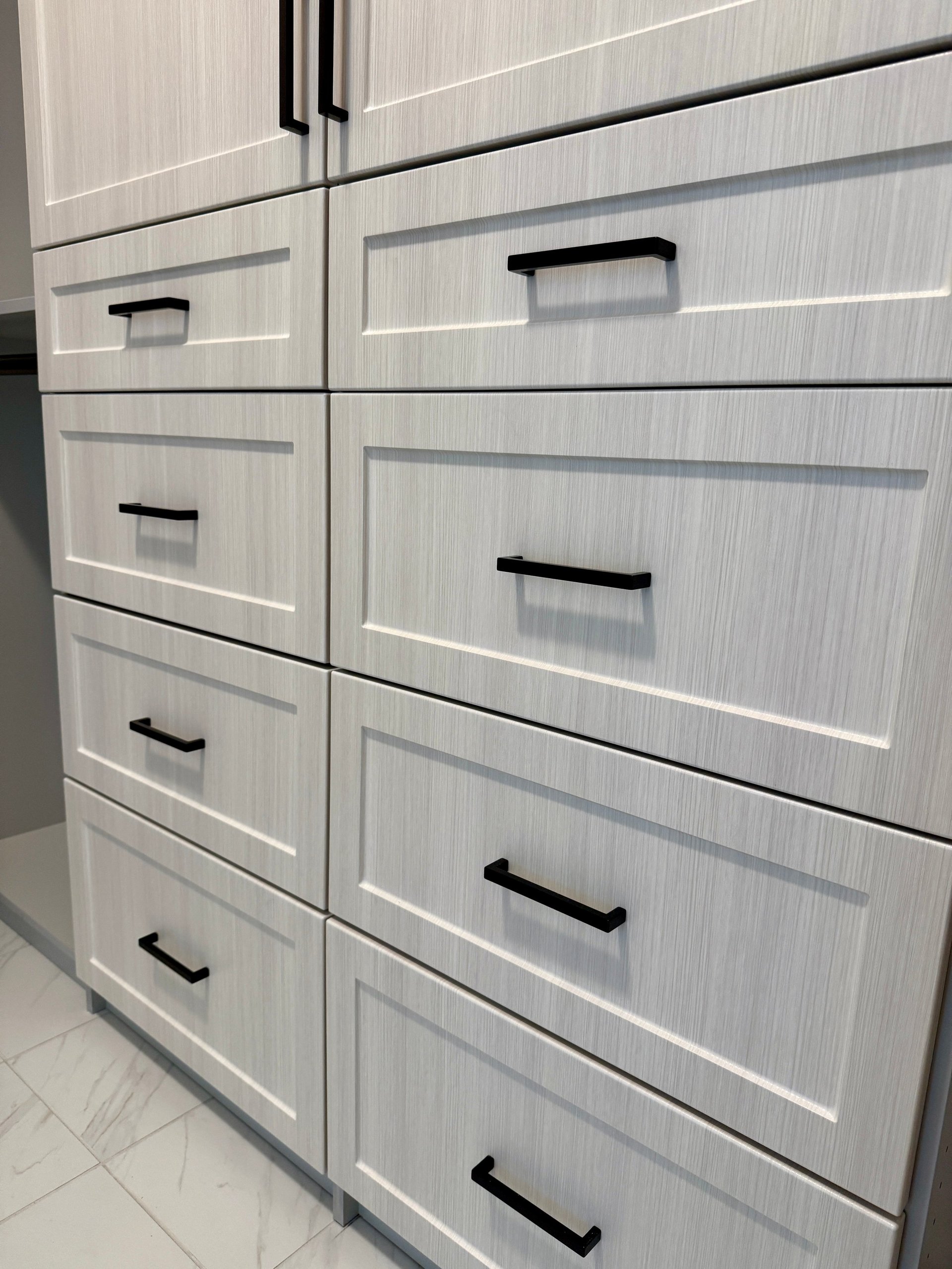 White wood grain cabinet with black metal handles arranged in a grid pattern of drawers and compartments